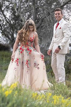 a young man and woman dressed in formal wear walking through tall grass with trees behind them