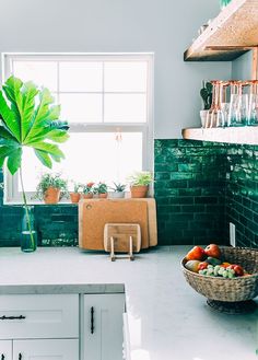 a kitchen with green tiles and plants in the window sill next to a basket of fruit