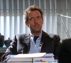 a man in a suit sitting at a desk with books and a lamp behind him