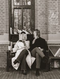 two people sitting on a bench with books in front of a brick building, one holding a book and the other reading