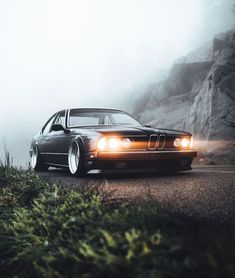 a black car driving down a road next to a lush green hillside on a foggy day