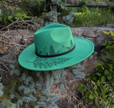 This hand burnt wide brim fedora makes a perfect addition to any nature lover's  wardrobe. The Kelly green material burns a gorgeous shade of emerald green allowing the design to stand out. I burnt pine trees in front of a mountain scene around the entire crown of the hat. Each mountain is intricately burned by hand with a pyrography machine using various tips to achieve depth and a rock-like texture.  On the right side brim of the hat I burnt a realistic fern. For a finishing touch, I burnt dis Artisan Style Fedora With Flat Brim For Outdoor, Custom Brimmed Fedora For Outdoor, Handmade Adjustable Fedora For Outdoor, Artisan Green Fedora Hat, Handmade Curved Brim Hat For Outdoors, Handmade Curved Brim Hat For Outdoor, Handmade Short Brim Hat For Outdoor, Handmade Flat Brim Hats For Outdoor, Custom Handmade Hats For Outdoors