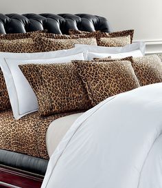 a bed covered in leopard print pillows and white sheets with black leather headboard behind it