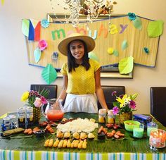 a woman wearing a hat standing behind a table with food on it and drinks in front of her