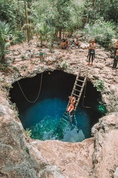 two people climbing up a ladder into a deep blue hole in the ground surrounded by trees