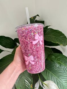 a hand holding a pink glittered cup with bows on the side and green leaves behind it