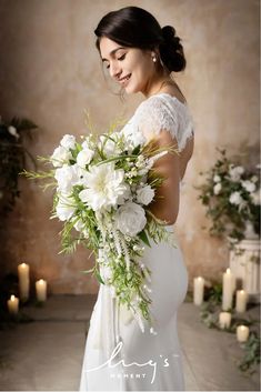 a woman in a white dress holding a bouquet of flowers and greenery with candles behind her