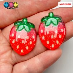 two red and green strawberry shaped glass cabochons in the palm of someone's hand