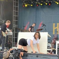 a woman in black shorts and white shirt standing on stage with guitar players behind her