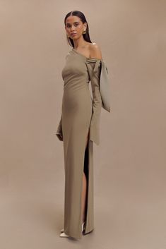 a woman in a long dress with an open shoulder and slited leg, standing against a