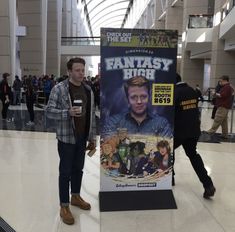 two men are standing next to a poster in an airport lobby, one is holding a drink