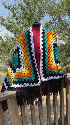 a colorful crocheted jacket is hanging on a wooden rail near a tree and fence