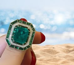 a close up of a person's hand holding an emerald and diamond ring on the beach