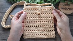 two hands holding a crocheted purse on top of a wooden table next to plants