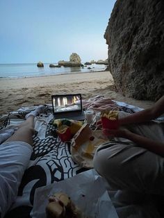 two people are sitting on the beach watching tv and eating food with their feet in the sand