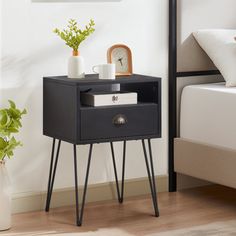 a nightstand with a clock on it next to a bed and plant in a vase