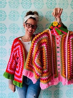 a woman holding up two crocheted sweaters on hangers in front of a wall