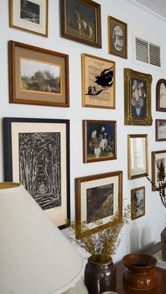 there are many framed pictures on the wall next to a lamp and vase with flowers in it