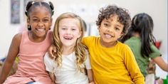 three young children sitting on the floor smiling at the camera with their arms around each other