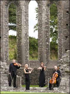 three people are playing violin in front of an old stone building with columns and arches