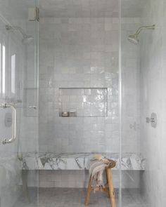 a walk in shower sitting next to a white tiled wall and floor with a wooden stool