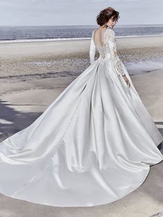 a woman in a white wedding dress standing on the beach with her back to the camera