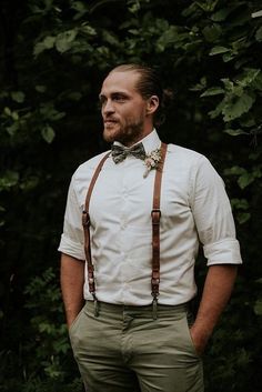 a man with a bow tie and suspenders standing in front of some green trees