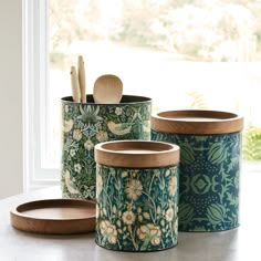 three canisters with wooden lids and spoons in front of a large window