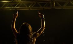 a woman standing in front of a crowd at a concert raising her arms to the sky