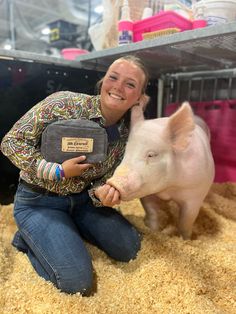 a woman kneeling down next to a small pig in a pen with hay on the floor