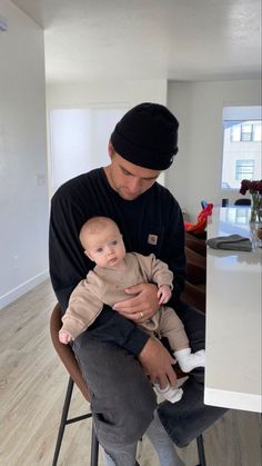 a man sitting in a chair holding a baby and looking at the camera while wearing a beanie