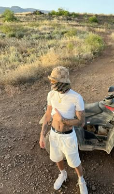 a man with tattoos standing next to a motorcycle in the dirt near grass and bushes