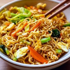 a bowl filled with noodles, vegetables and chopsticks on top of a wooden table