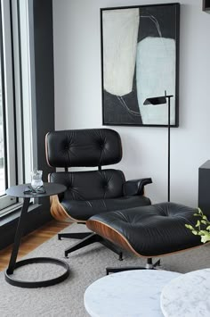 an eames chair and ottoman in front of a window with a painting on the wall