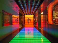 a brightly lit hallway with people walking on the floor and colorful lights shining down from the ceiling