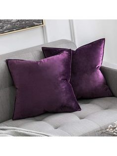 two purple pillows sitting on top of a gray couch next to a white vase with flowers