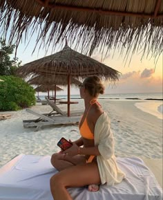 a woman sitting on top of a white bed under a straw hut next to the ocean