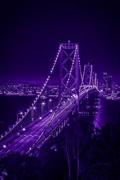 the bay bridge in san francisco is lit up with purple lights at night, as well as city lights