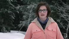 a woman wearing glasses standing in front of snow covered trees and looking at the camera