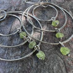 "I have carefully wrapped a set of genuine raw Peridot gemstones in sterling silver wire.  The small gems dangle from sterling silver hoops I have formed, soldered, and textured by hand.  Earrings dangle from handmade sterling silver earwires.  The silver has been oxidized and polished to create a patina finish. Peridot: About 4-5mm Total Earring Length (Including Earwire): About 2.75\"  The largest hoop measures just over 1.5 inches in diameter. Total Earring Weight: About 3.5 grams per one earring Be sure to see all photos and video for size and color reference. Wise Handmade Jewelry is an eco-conscious company.  Jewelry will be carefully packaged in earth-friendly and sustainable materials. For more information about Wise Handmade Jewelry, visit wisehandmade.com" Green Small Hoop Sterling Silver Jewelry, Green Sterling Silver Small Hoop Jewelry, Fusion Style Sterling Silver Hoop Earrings As Gift, Hand Wrapped Dangle Jewelry In Silver Plated Wire, Hand Wrapped Green Sterling Silver Jewelry, Hand Wrapped Silver Plated Dangle Jewelry, Handmade Peridot Dangle Jewelry, Wire Wrapped Small Hoop Sterling Silver Earrings, Silver Dangle Hoop Earrings With Natural Stones