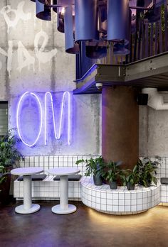 two white benches sitting in front of a neon sign on the wall above them are plants and potted plants