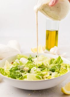 someone pouring dressing into a salad in a white bowl with lettuce and lemon wedges