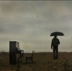 a man holding an umbrella standing next to a piano and chair in the middle of a field