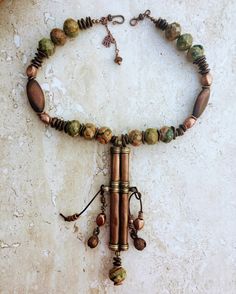 This necklace features an authentic vintage Tibetan Needle Box re-purposed into a piece of unique statement jewelry. I did not envision the piece to be overly colorful so I teamed up the copper patina on the pendant with the green, mustard brown and copper shades of the Kabamba Rhyolite Jasper stone beads. These beads are chunky and heavy - each one being 20mm wide so it gives the necklace a lot of oomph and character. The wood and copper accents add texture to the necklace and the Bronzite stone pebble beads tie the colors and ground the entire piece. I retained the authentic bells that came with the pendant (they hung on very worn out and frayed leather cords on either sides of the needle box) and restrung them in cow-hide leather cords along with copper accents to give the necklace move Unique One-of-a-kind Copper Necklace, Vintage Bronze Necklaces With Natural Stones, Rustic Hand Wrapped Jewelry For Festivals, Vintage Brass Necklaces For Rituals, Bohemian Brass Necklace With Patina, One Of A Kind Bronze Necklace For Festival, Bohemian Hand Wrapped Brass Necklace, Unique Bronze Necklace For Festivals, Rustic Patina Jewelry For Festival