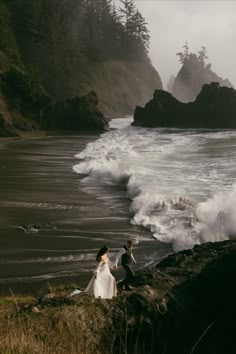 a man and woman standing on top of a cliff next to the ocean with crashing waves