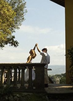 two people are holding hands on a porch with a dog in the foreground and trees behind them