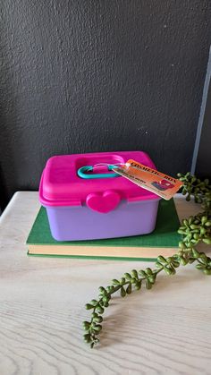 a pink and purple lunch box sitting on top of a green book next to a plant