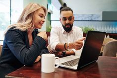 two people sitting at a table looking at a laptop computer and talking to each other