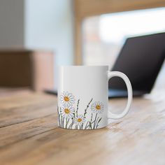 a white coffee mug with daisies on it sitting on a table next to a laptop