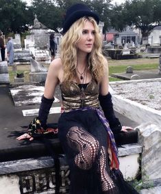 a woman sitting on top of a stone wall next to a cemetery wearing a black hat